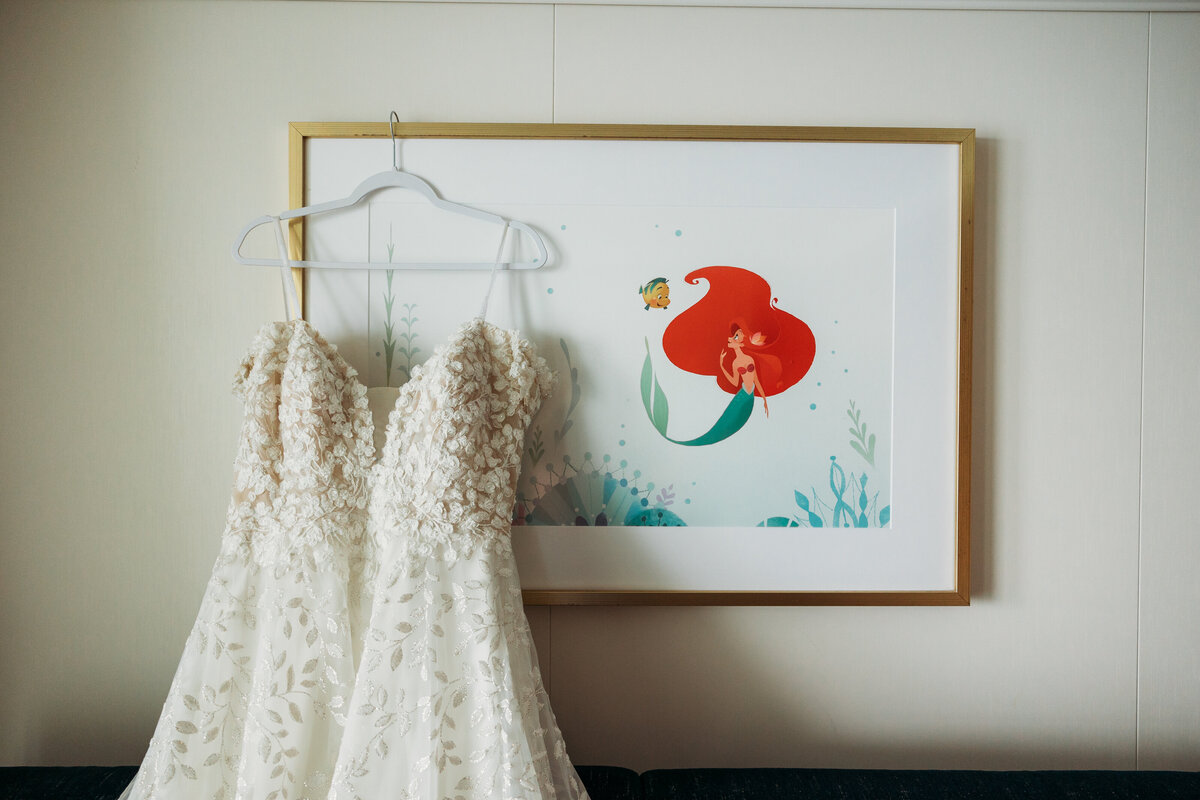 Floral applique bridal gown hanging on a white hanger in front of Ariel and Flounder artwork aboard the Disney Wish