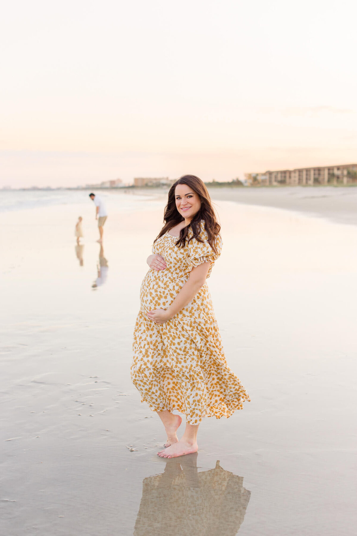 Pregnant mother stands on the beach holding her belly while her husband and young toddler girl playing in the background at sunset