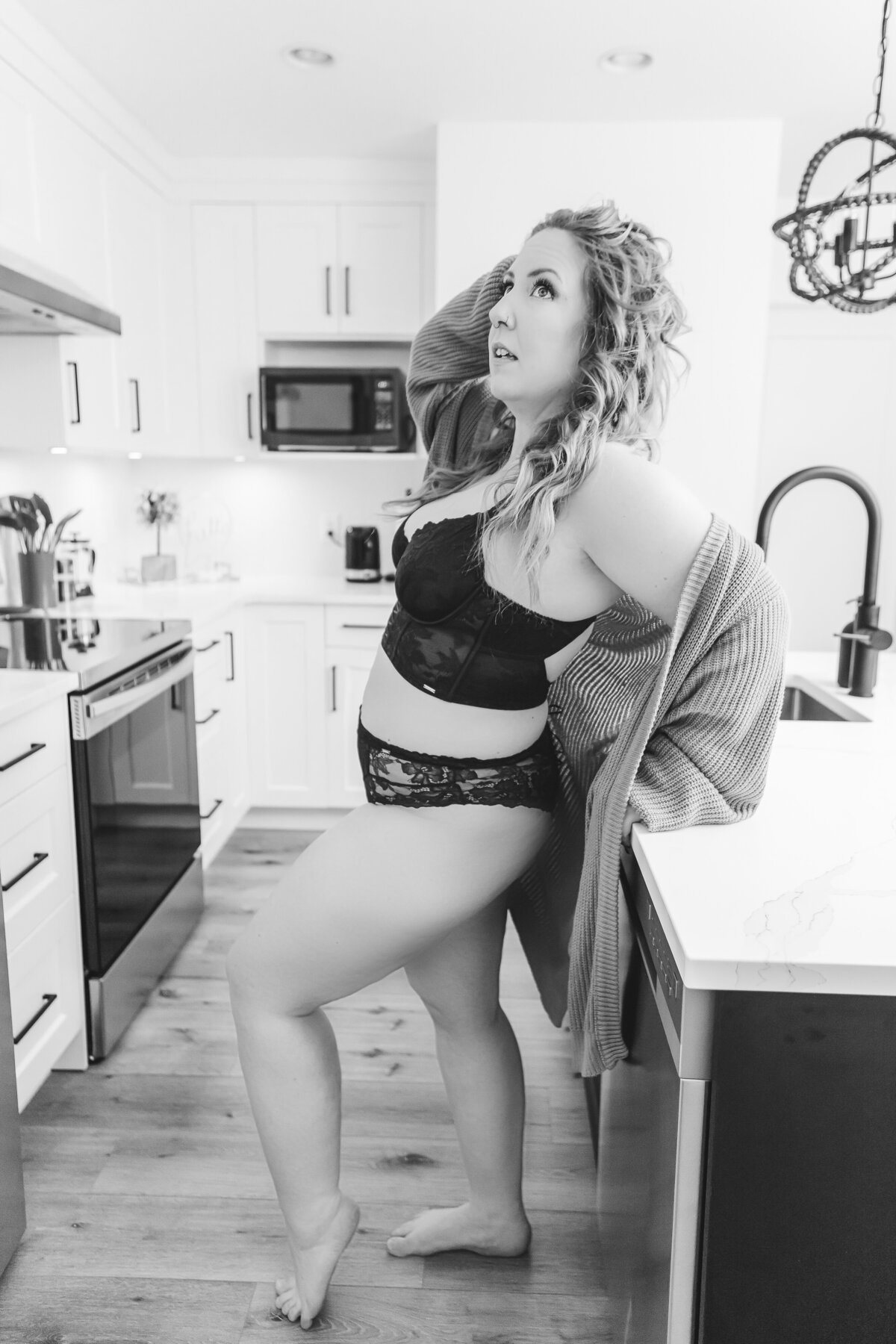A woman wearing black lingerie and a mustard cardigan, standing confidently in a modern kitchen.