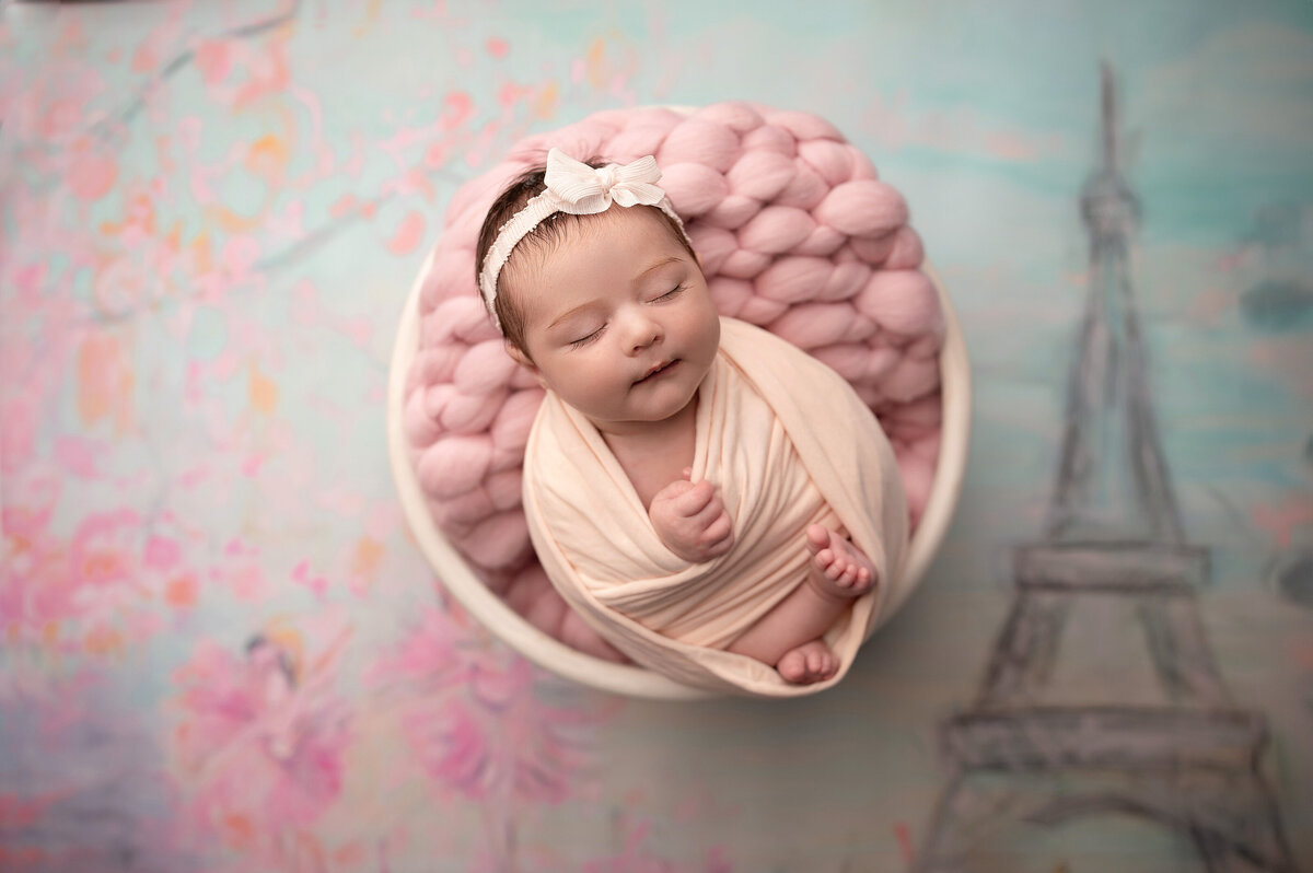 sleeping two week old newborn baby girl wrapped with hands and feet sticking out posed in round white bowl with pink layer over a background with the eiffel tower