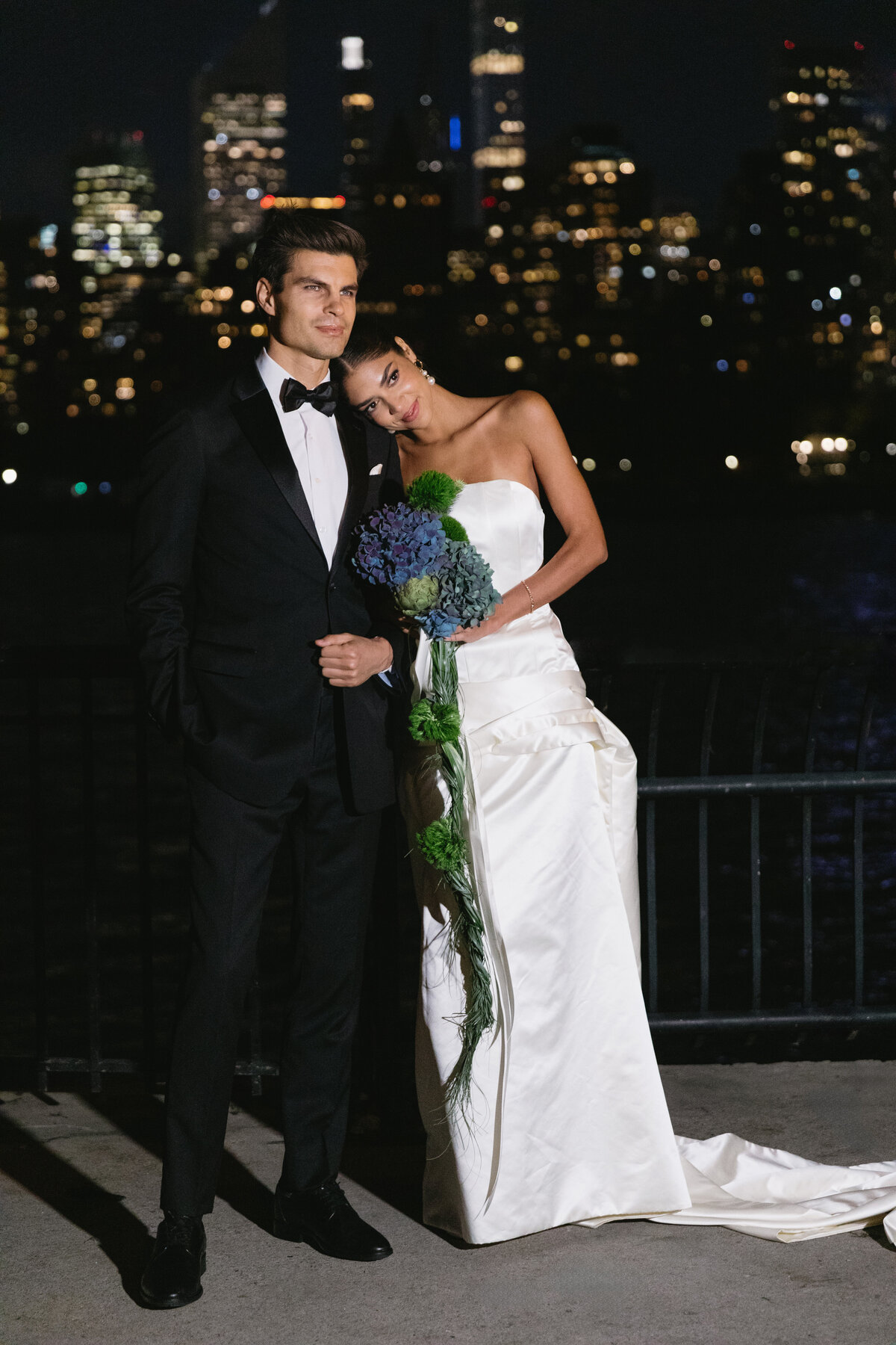 Bride and groom posing at night with city lights in the background and a hydrangea bouquet.