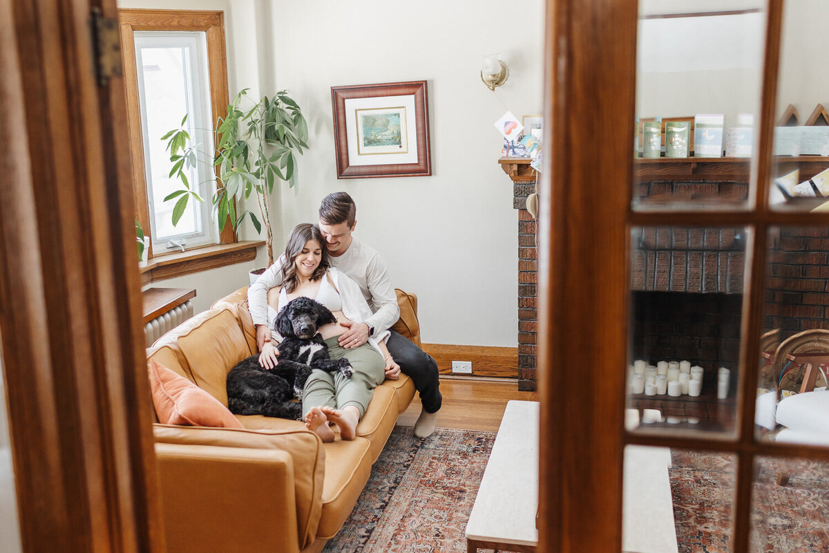 Expecting couple sitting on their brown leather couch with their adorable black dog