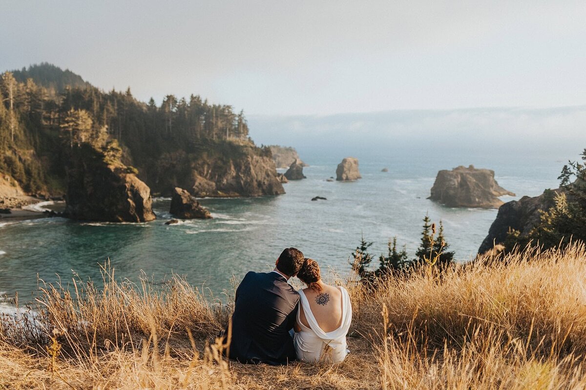 A couple sits on a cliff edge in wedding attire overlooking the Oregon Coast