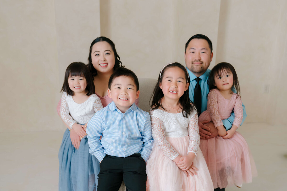 Parents and four children sit closely together in a timeless studio family portrait with soft beige tones.
