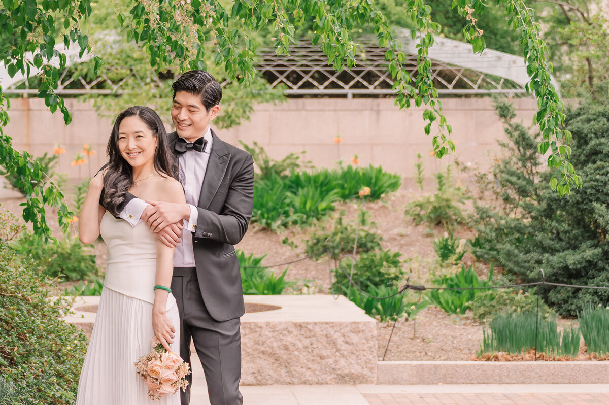 An Asian groom in a black tux snuggling his fiance around the shoulder in a white dress underneath greenery in a Washington DC park at sunrise as they enjoy their destination wedding photos by JoLynn Photography