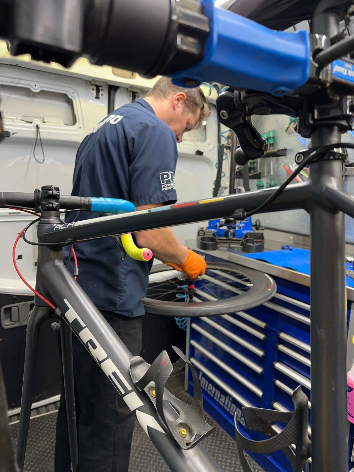 A mechanic wearing orange gloves works on a bicycle wheel inside a workshop van. A bicycle frame is in the foreground, and various tools are visible on a blue workbench in the background