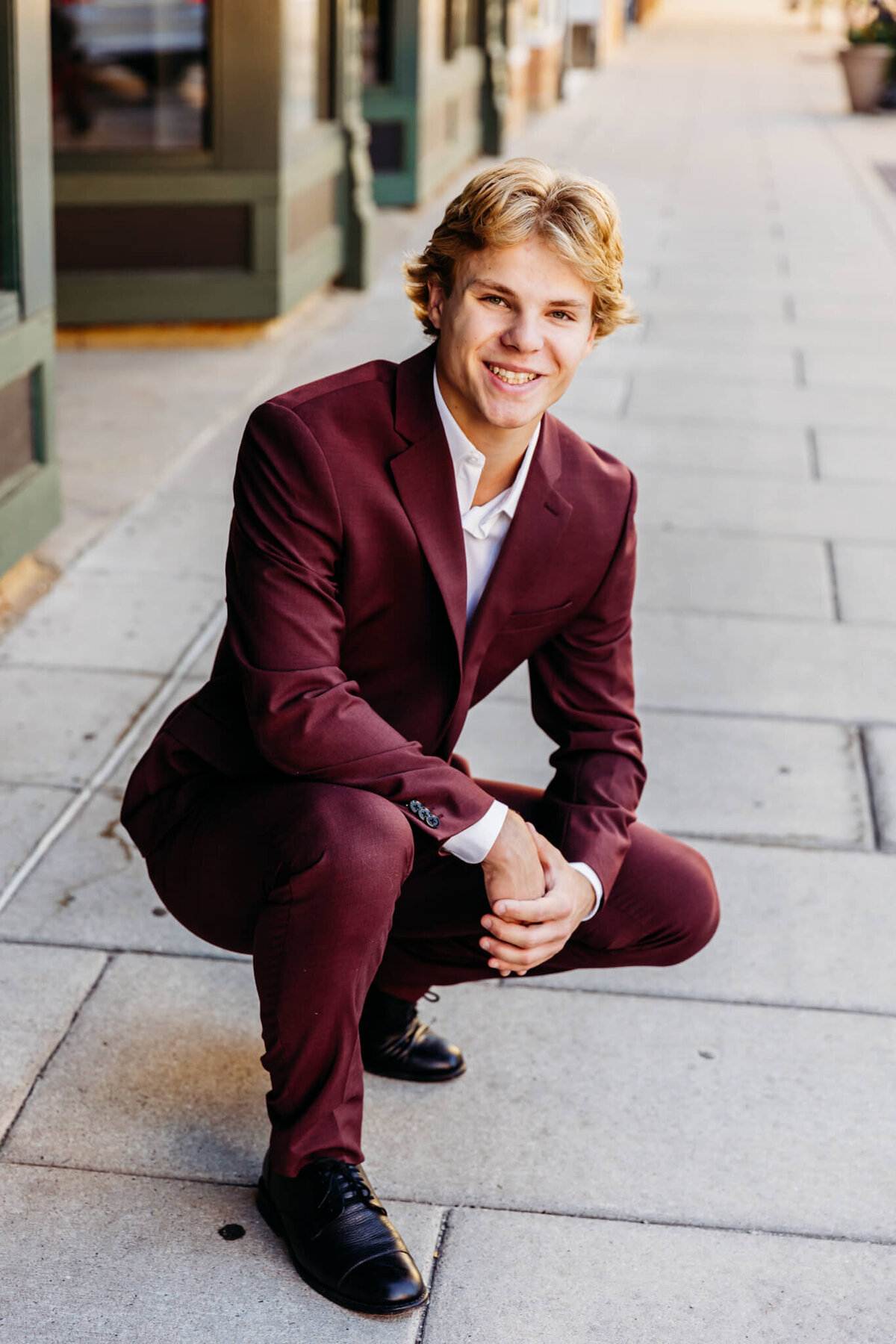 male high schooler kneeling on a sidewalk and smiling as he holds his hands together captured by Ashely Kalbus Photography