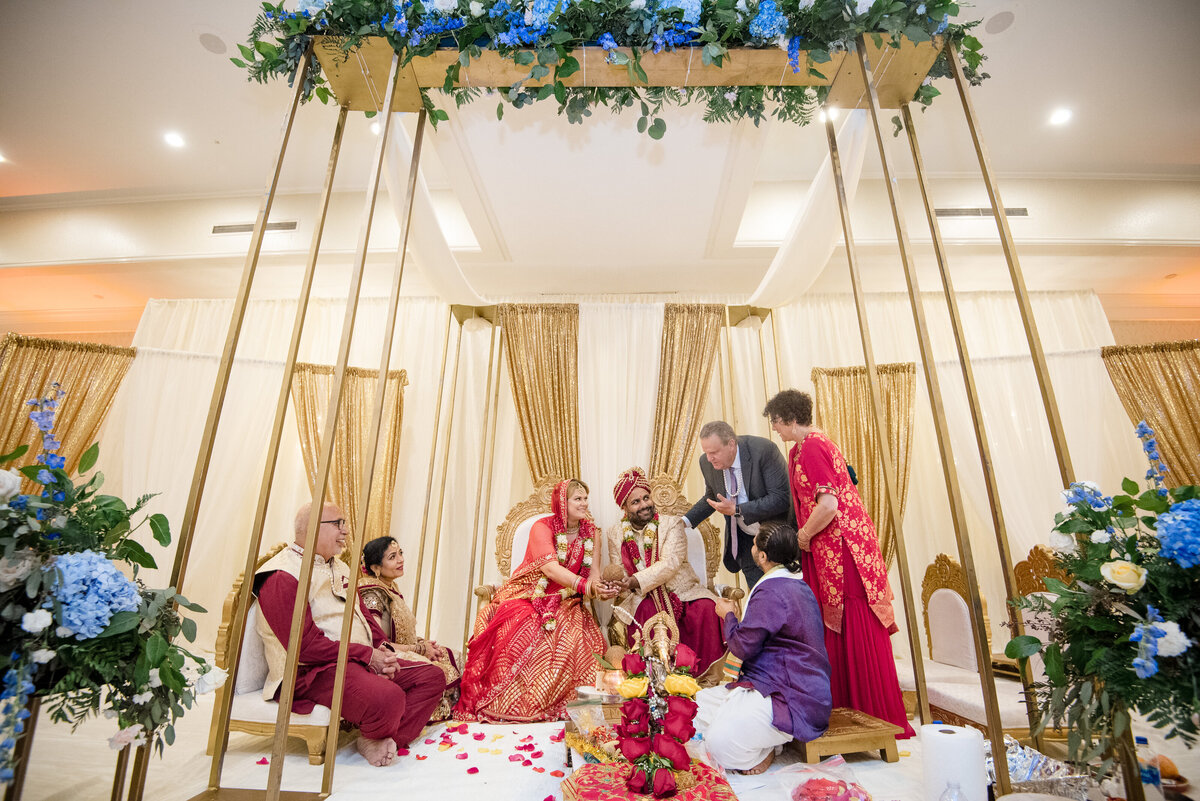 Traditional Indian wedding ceremony at Le Méridien Charlotte. The bride and groom, dressed in red and gold attire, sit together on a decorated mandap surrounded by family and friends during sacred rituals. Captured by a Charlotte Indian wedding photographer specializing in cultural and candid wedding moments.