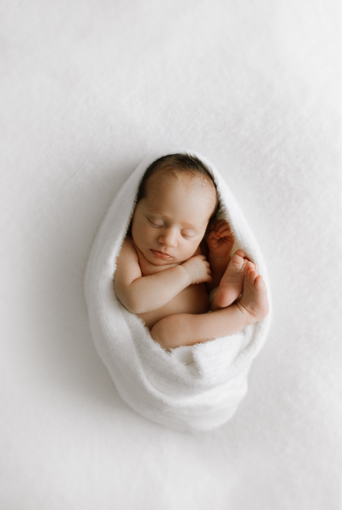 A newborn baby peacefully sleeps wrapped snugly in a soft white blanket, posed in a gentle curl against a creamy white backdrop. The minimal setup draws focus to the baby’s delicate features, with toes tucked close and hands near the face. Captured in a Sydney newborn studio using diffused artificial light.