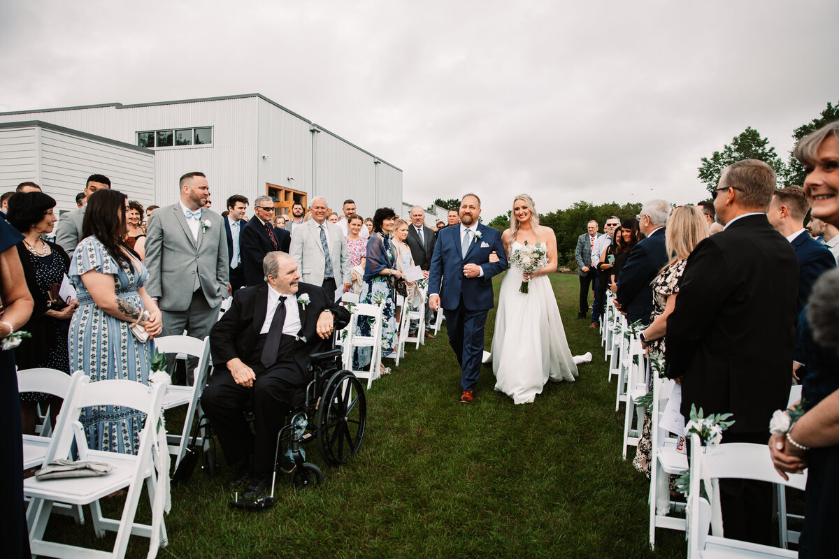 A bride walks down the aisle holding dad's arm to her outdoor Aspen Sky Winery and Distillery wedding