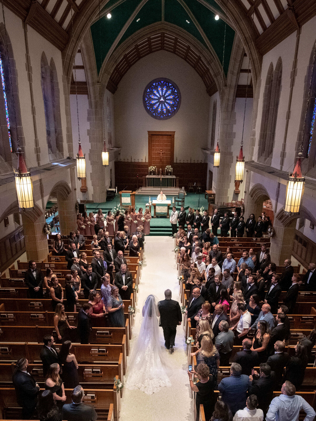 A bride in a long white gown walks down the aisle with a man, likely her father, in a large church filled with seated guests. The ornate interior features high ceilings, stained glass windows, and a circular stained glass piece above the altar.