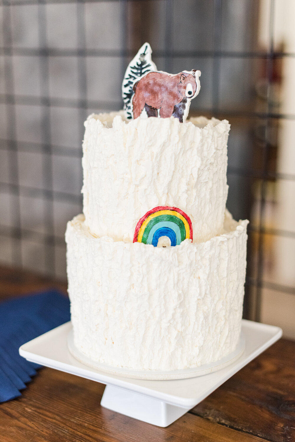 Two-tier woodland baby shower cake with buttercream textured to resemble tree bark, topped with a whimsical bear and tree, and featuring a hand-painted rainbow on the side.