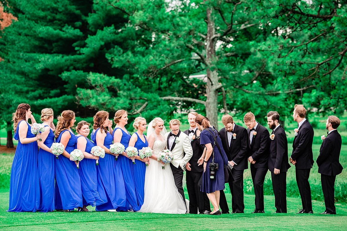 Photographer chatting with groom during wedding party photos on the front lawn of the Country Club, bridesmaids are wearing a blue full length dress and groomsmen are in classic black tuxedos