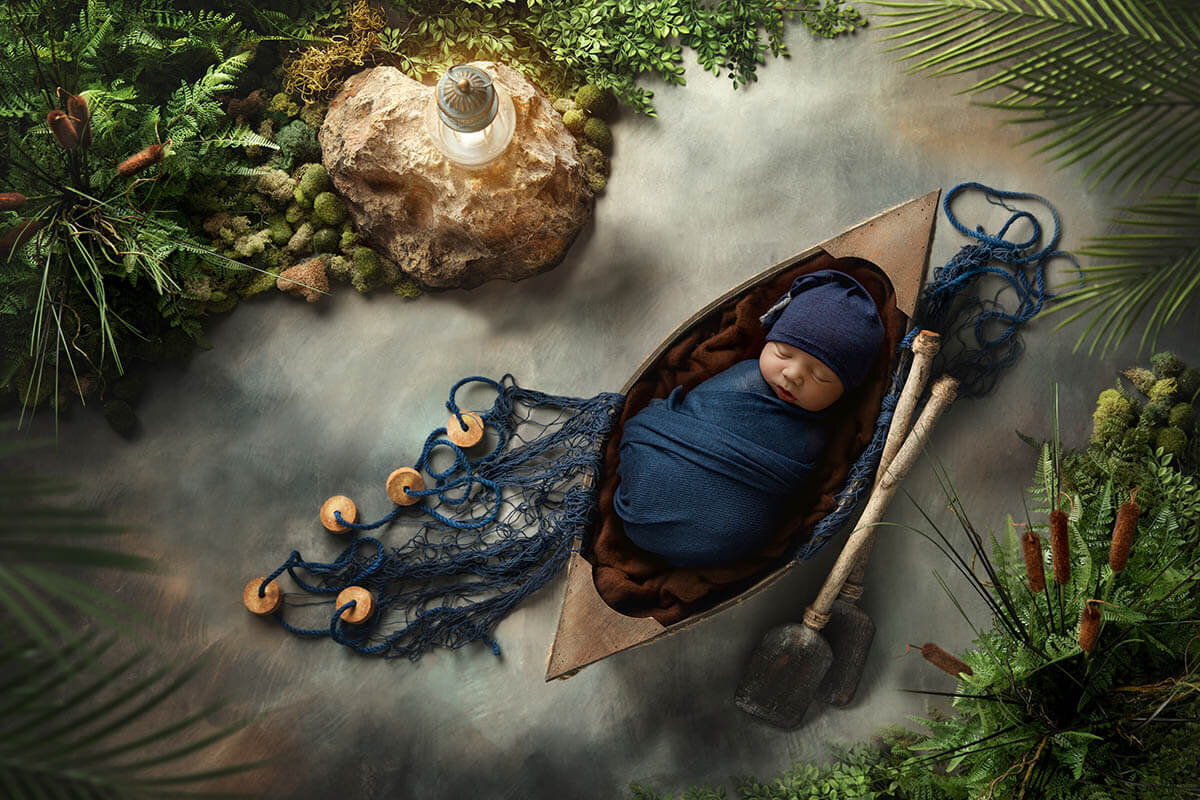 Sleeping infant boy swaddled in navy rests in a canoe surrounded by greenery, paddles, and netting.