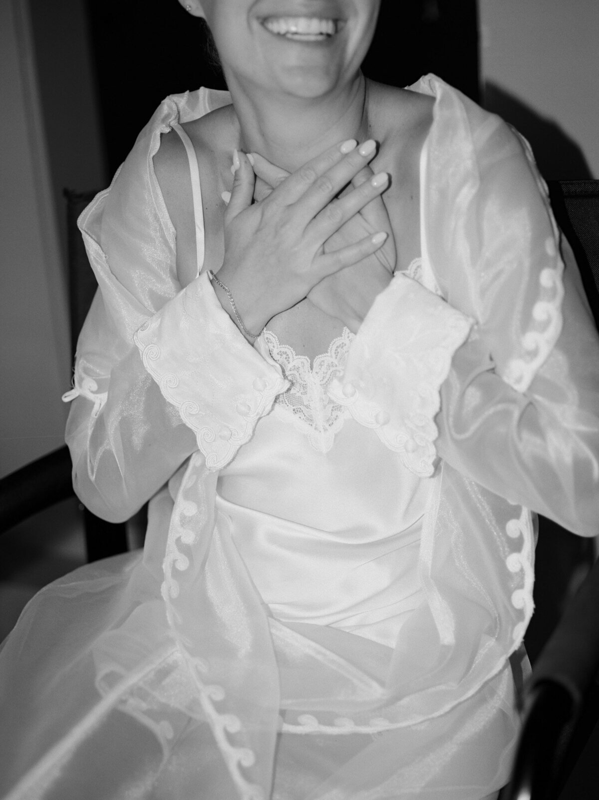 A bride laughs with her hands over her heart in a silk & lace robe while having makeup done for her Detroit wedding