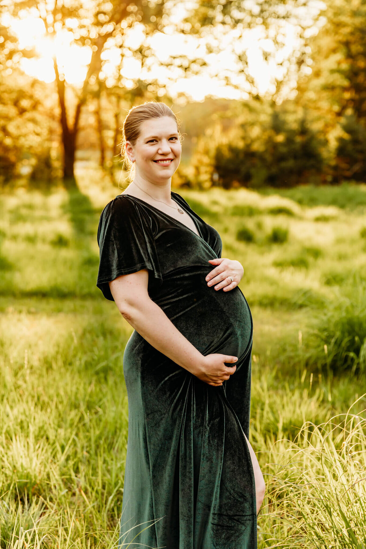 stunning mom to be in a green velvet dress holding her baby bump in a field.