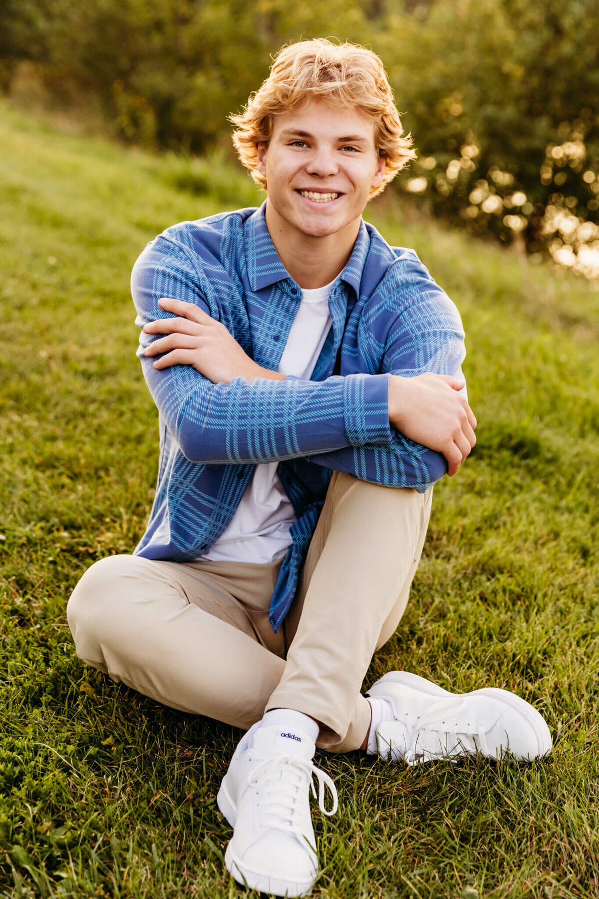 young man sitting with his legs crossed and his arms resting on his knee as he smiles for his senior photos.