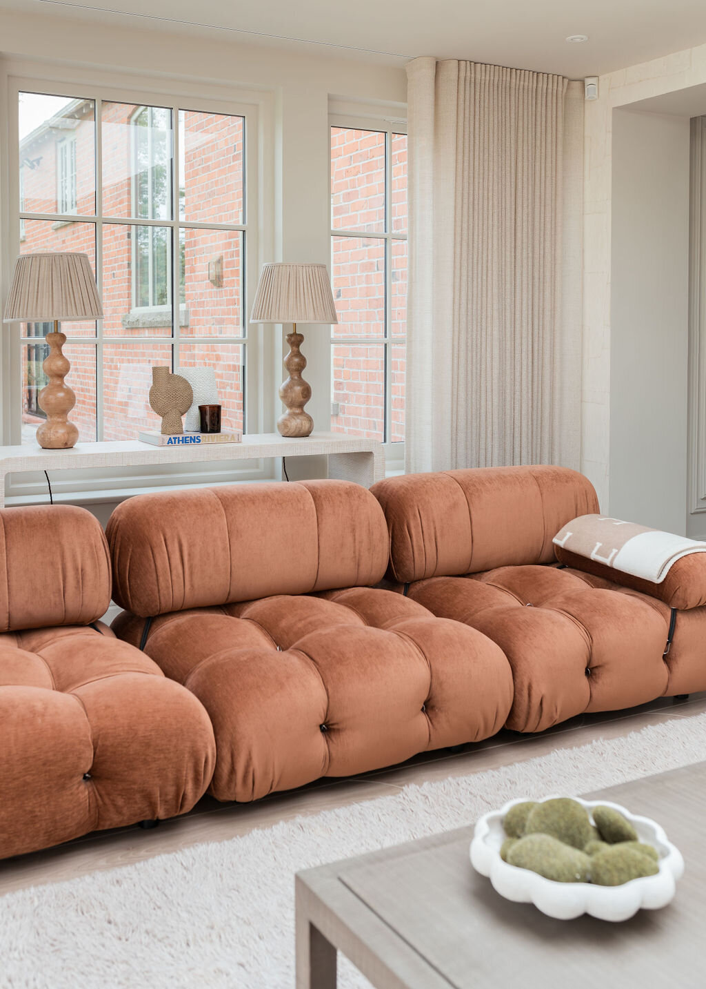 A cozy living room with large windows features a plush, rust orange tufted sofa, a light-colored rug, and a wooden coffee table adorned with decorative items and books. Two large, neutral lamps sit on a console table behind the sofa.