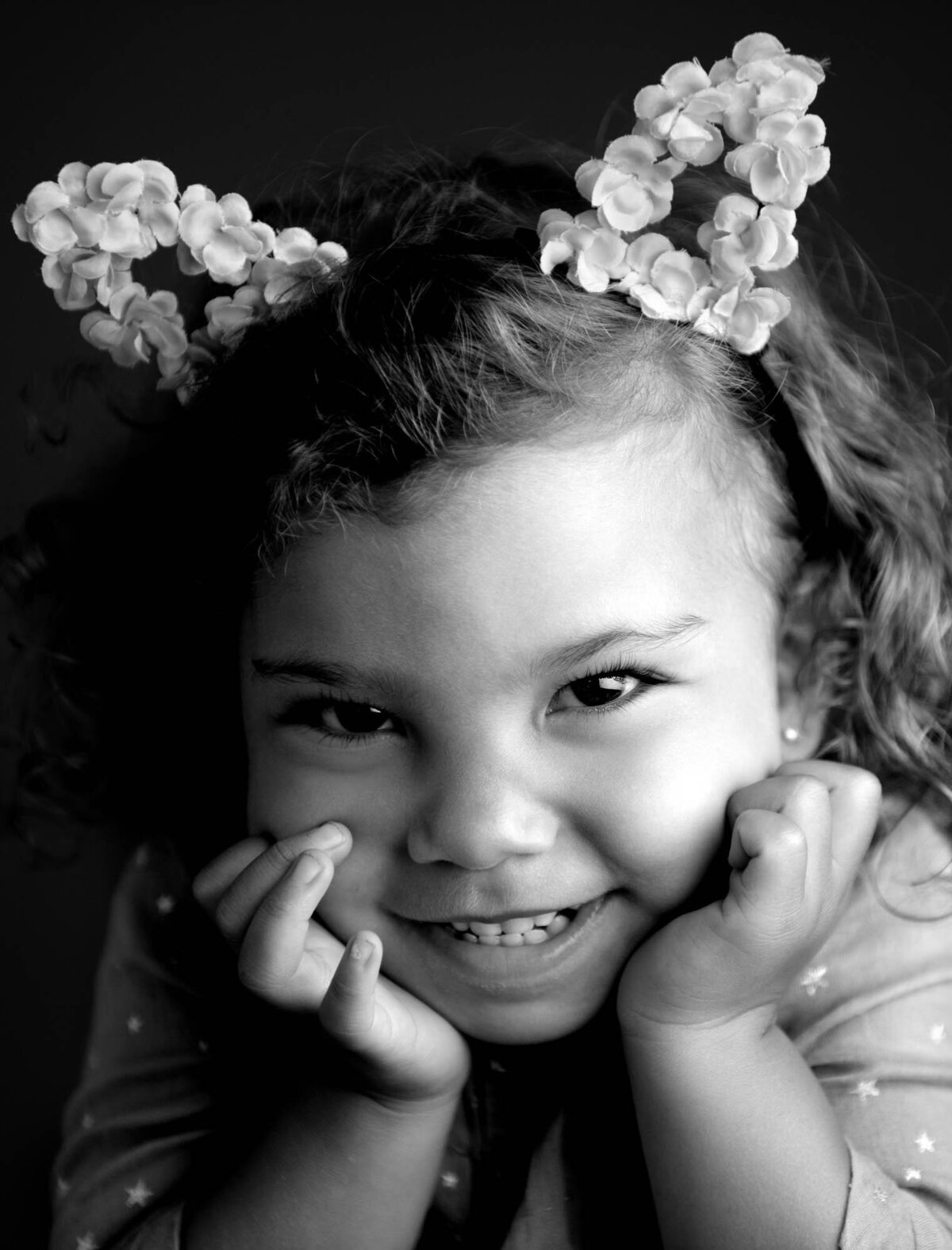 Smiling child with curly hair and flower-bedecked cat ear headband. Black and white close-up portrait creating a playful, joyful mood.