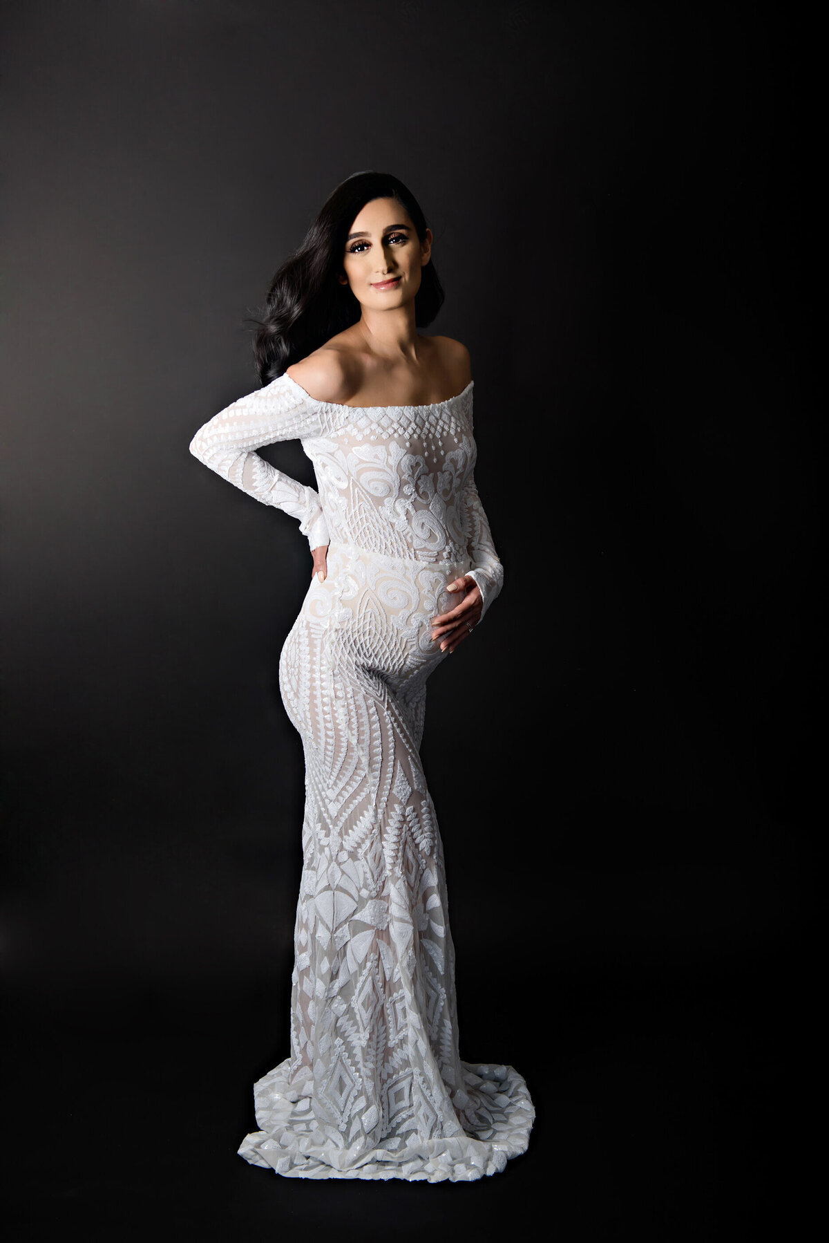 Brown haired pregnant woman wearing a white dress against a black background holding her belly for her in-studio Burlington, ON maternity photography session