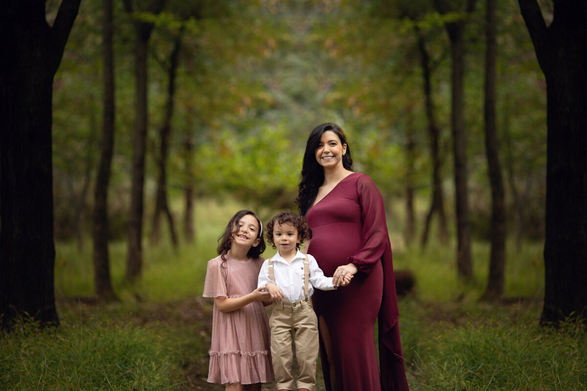 A pregnant mother holds hands with her toddler daughter and son while walking through a forest path