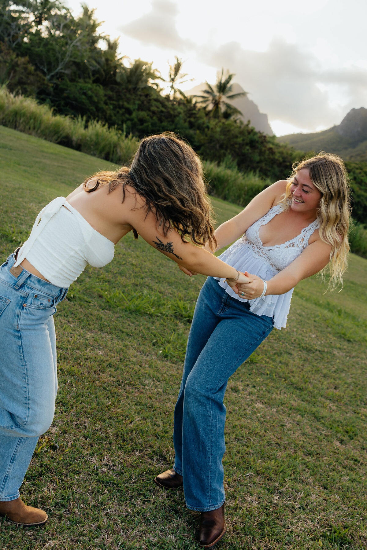 Two best friends laughing and holding hands while spinning around in a grassy field with mountain views in Oʻahu.