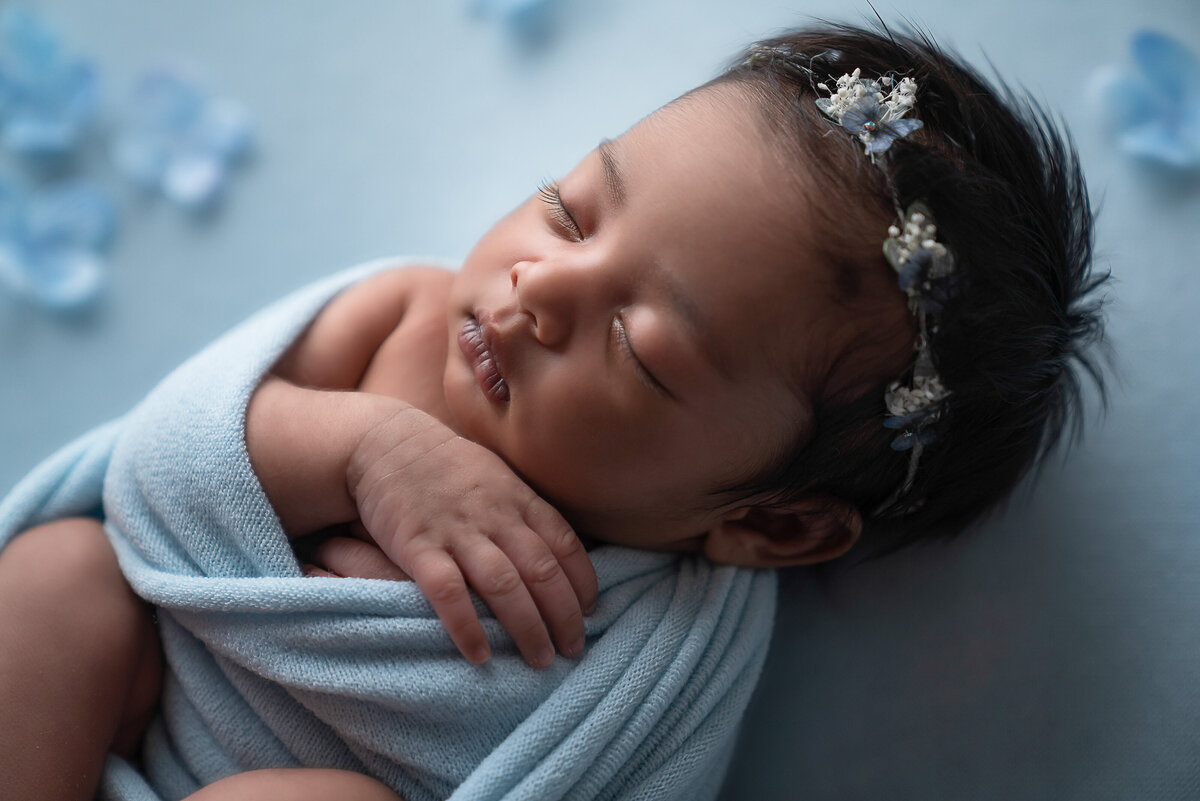 Baby girl swaddled in blue and waring a blue headband is sleeping with her feed and hands folded and exposed for newborn photoshoot. She is surrounded by blue flowers. Captured by Bryn Mwar Philadelphia and Main Line photographer Liza Nicole.