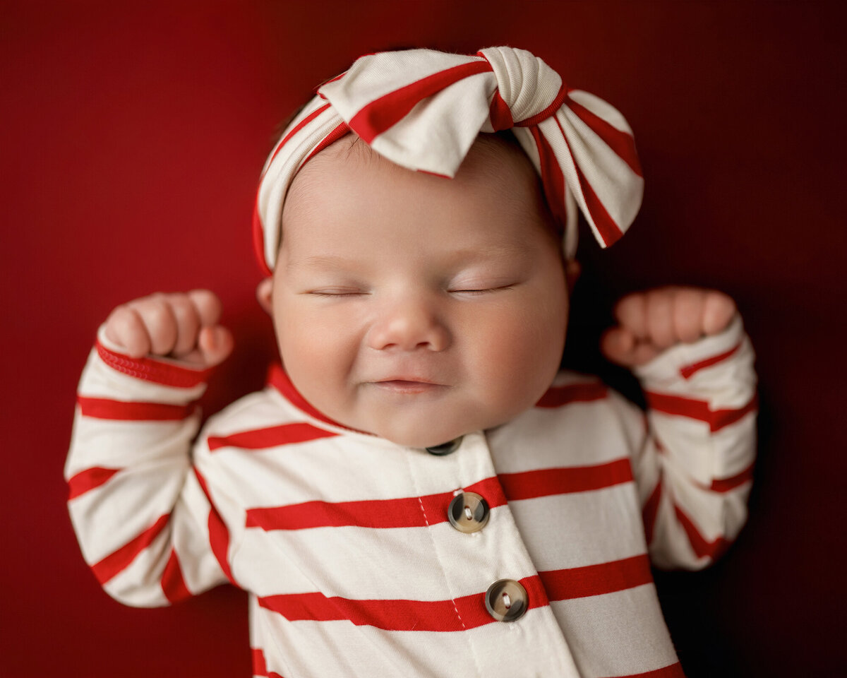 A baby wears a christmas stripe onesie on red bed with matching bow