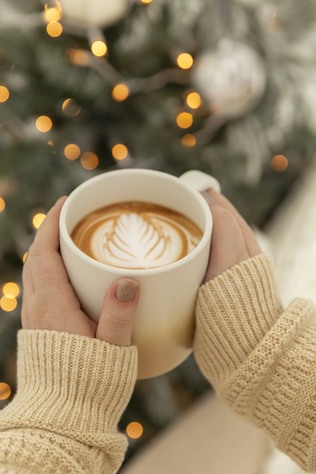 A pair of women's hands in a comfy sweater holding a large coffee mug with late art Infront of holiday lights.