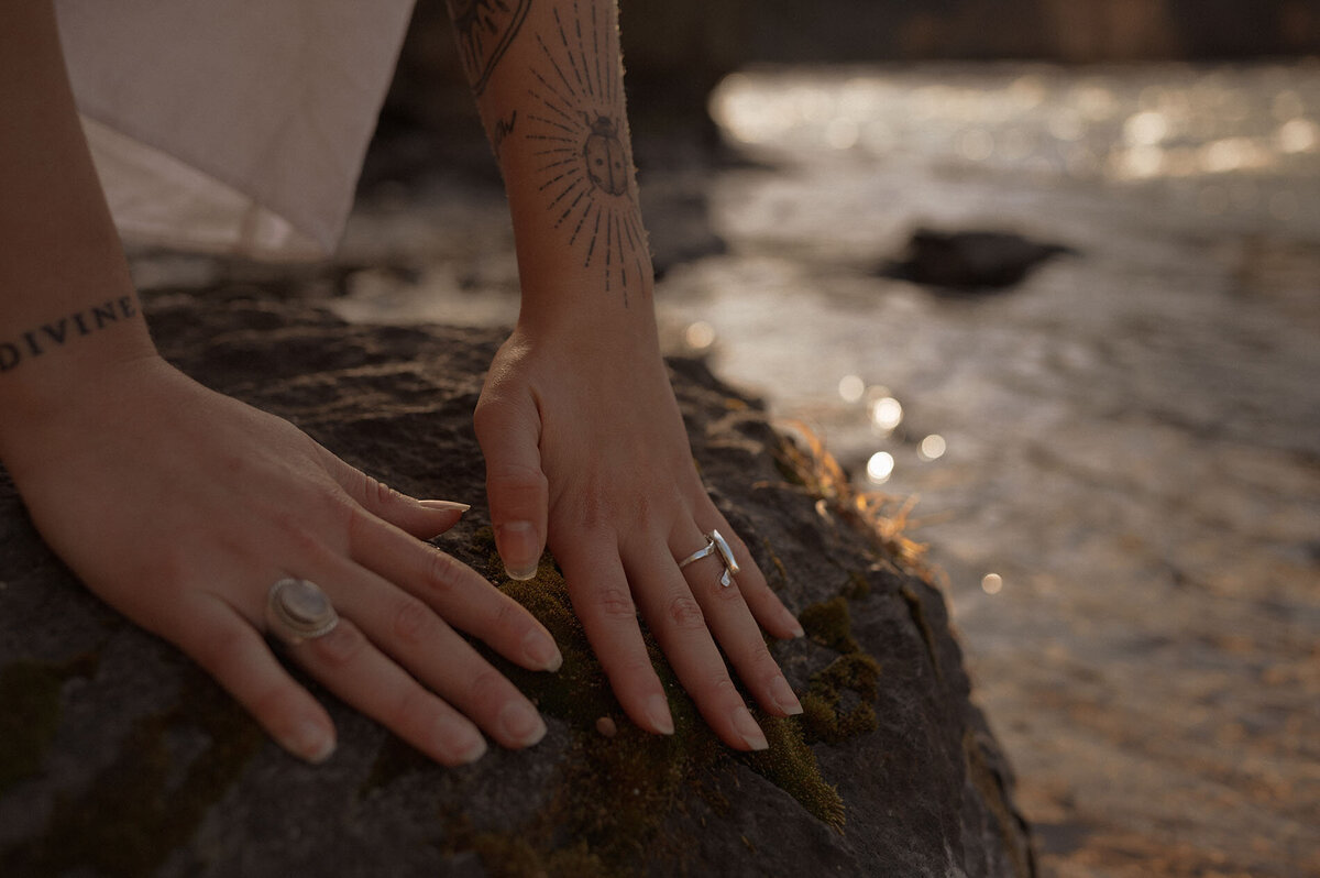 woman with tattoos and rings touches a rock near the river in Oregon