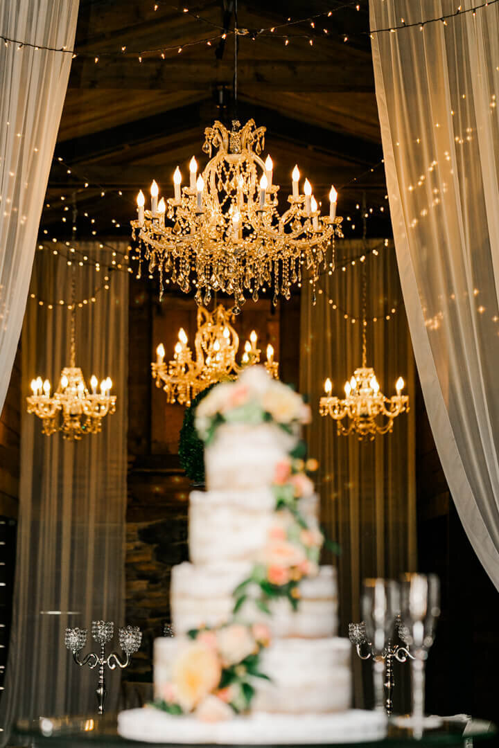 The bridal cake is shown in the foreground with warm lighted chandeliers lighting the room behind it.