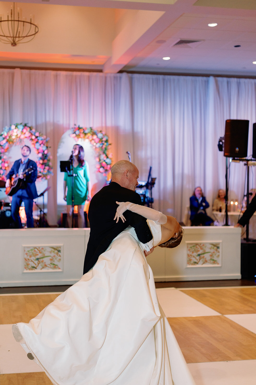 Bride in a white gown is dipped back gracefully by her father during their dance at the wedding reception, with a live band playing in the background and floral arches behind the stage.