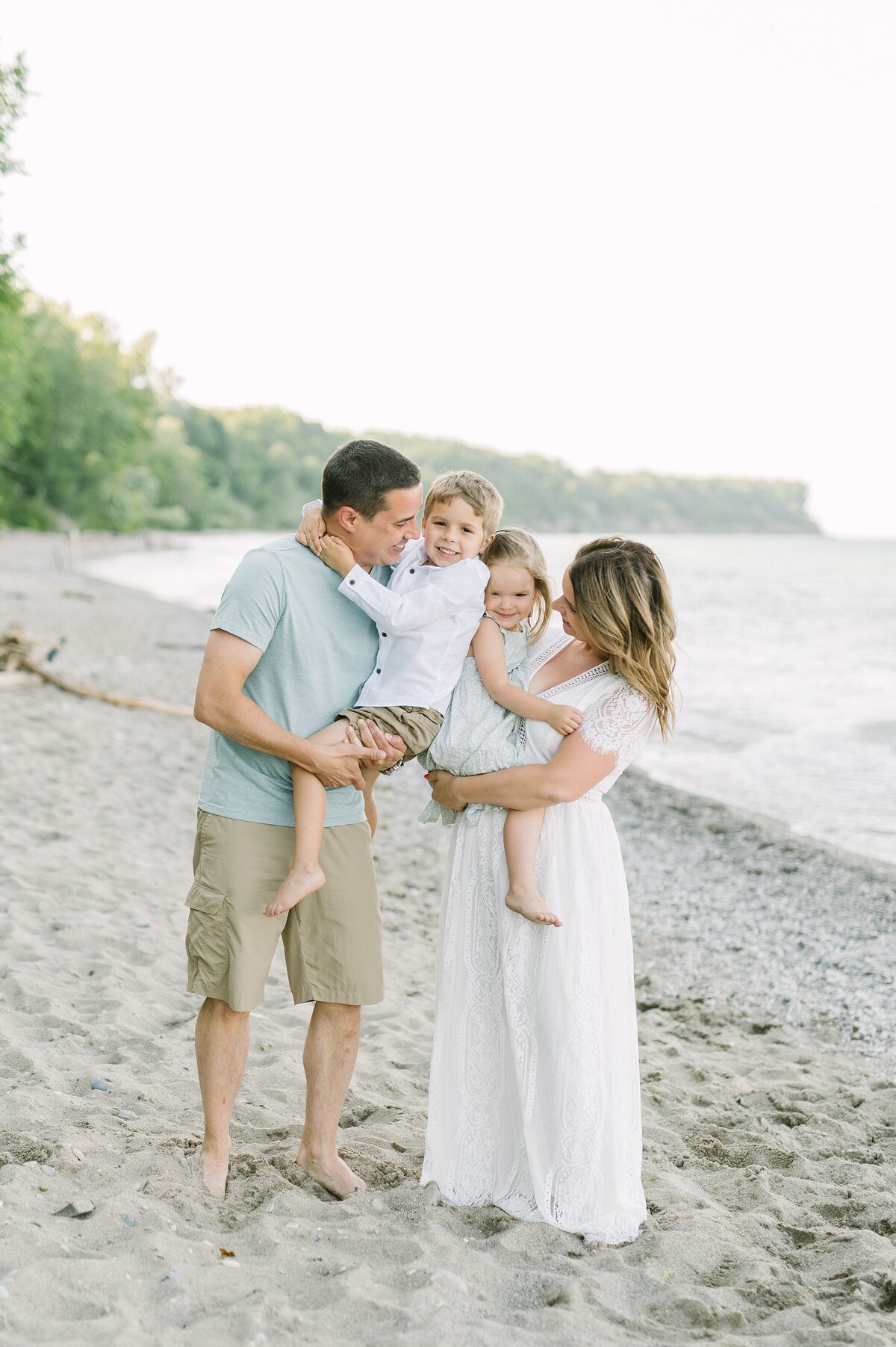 Mom and dad are each holding a kid and smiling toward the children on the beach