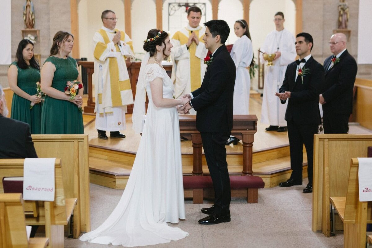 A bride and groom holding hands and their bridal party stand in front of an altar during their wedding ceremony.