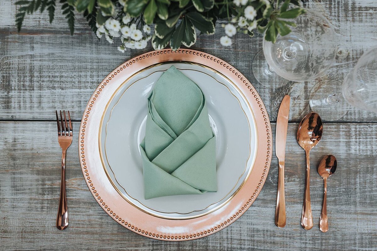 A copper charger with a white plate and copper flatware sitting on a gray farm table. Sage fancy folded napkin sitting in the center of the plate with a green and white centerpiece.