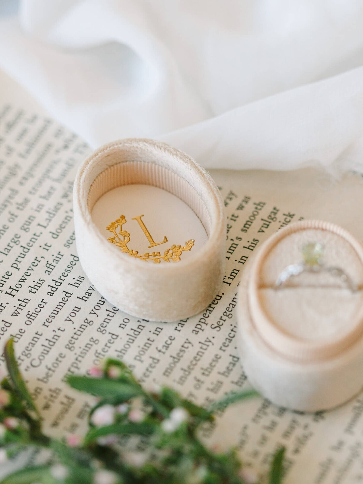 Velvet ring box on a book page, adorned with an "L" and floral design. An engagement ring sits beside it.