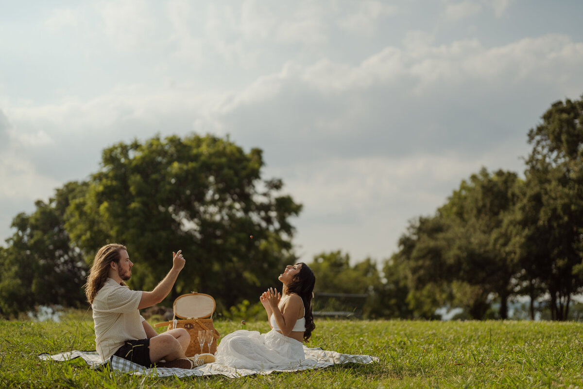A joyful spring engagement photo featuring a couple enjoying a playful picnic in the park near White Rock Lake in Dallas. Capturing laughter and lighthearted moments, this image highlights the natural charm and romance of outdoor sessions in Dallas and Fort Worth, Texas.