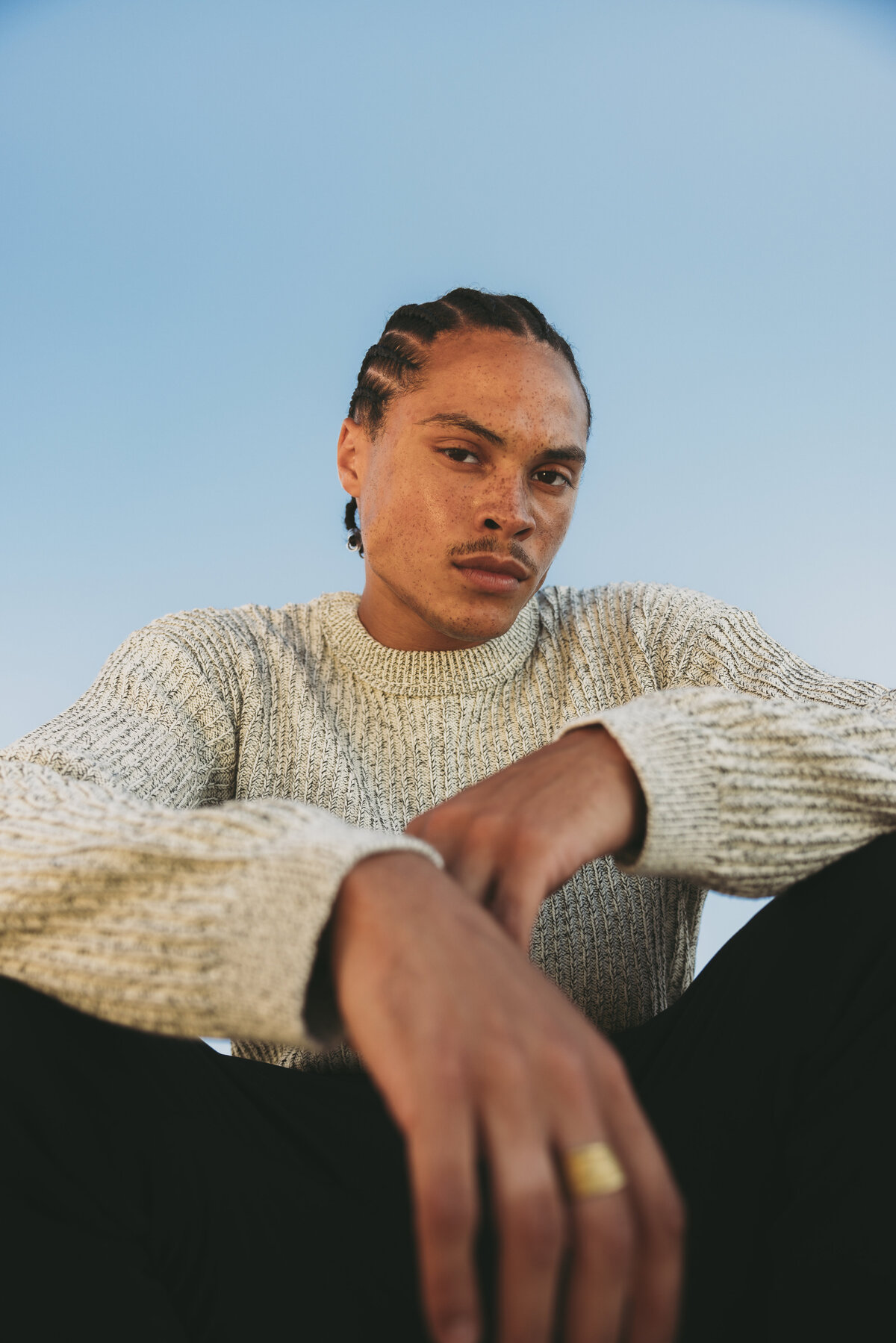 “Close-up portrait of Timothe wearing a textured beige sweater and gold ring, set against a clear blue sky in Cape Town.