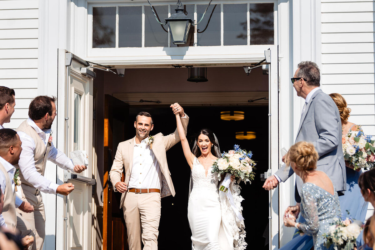 Newlyweds excitedly exiting a church, holding hands up in celebration, with guests and bridesmaids on either side cheering.
