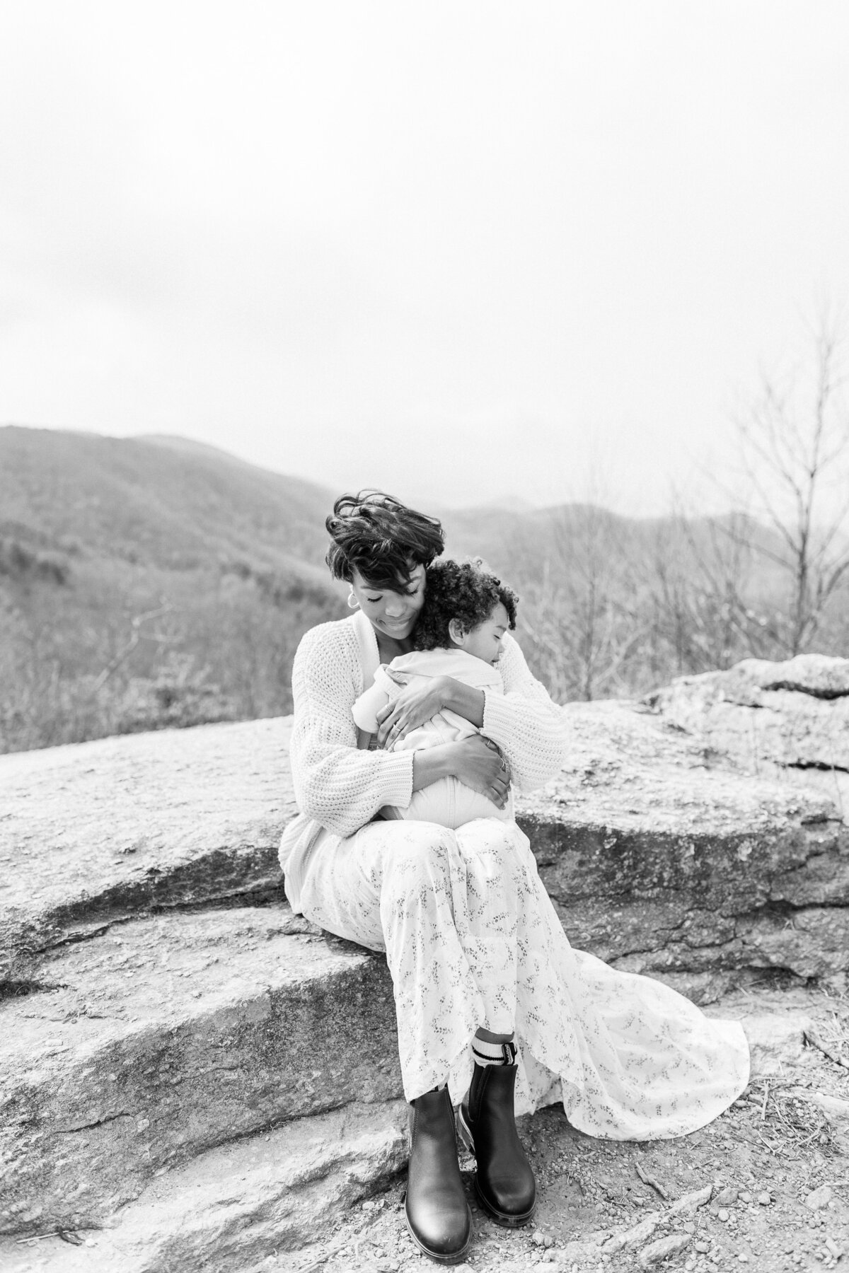 Black and white photo of mother seated with toddler son on a rock during a Kansas City newborn session.