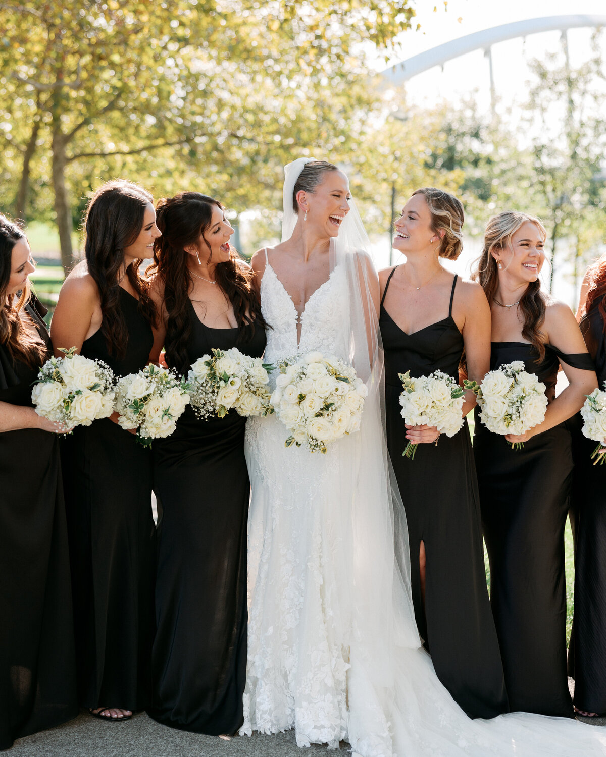 Bride and her bridesmaids laugh together holding white bouquets and wearing black gowns, full of joy and natural light—captured by a Philadelphia wedding photographer.
