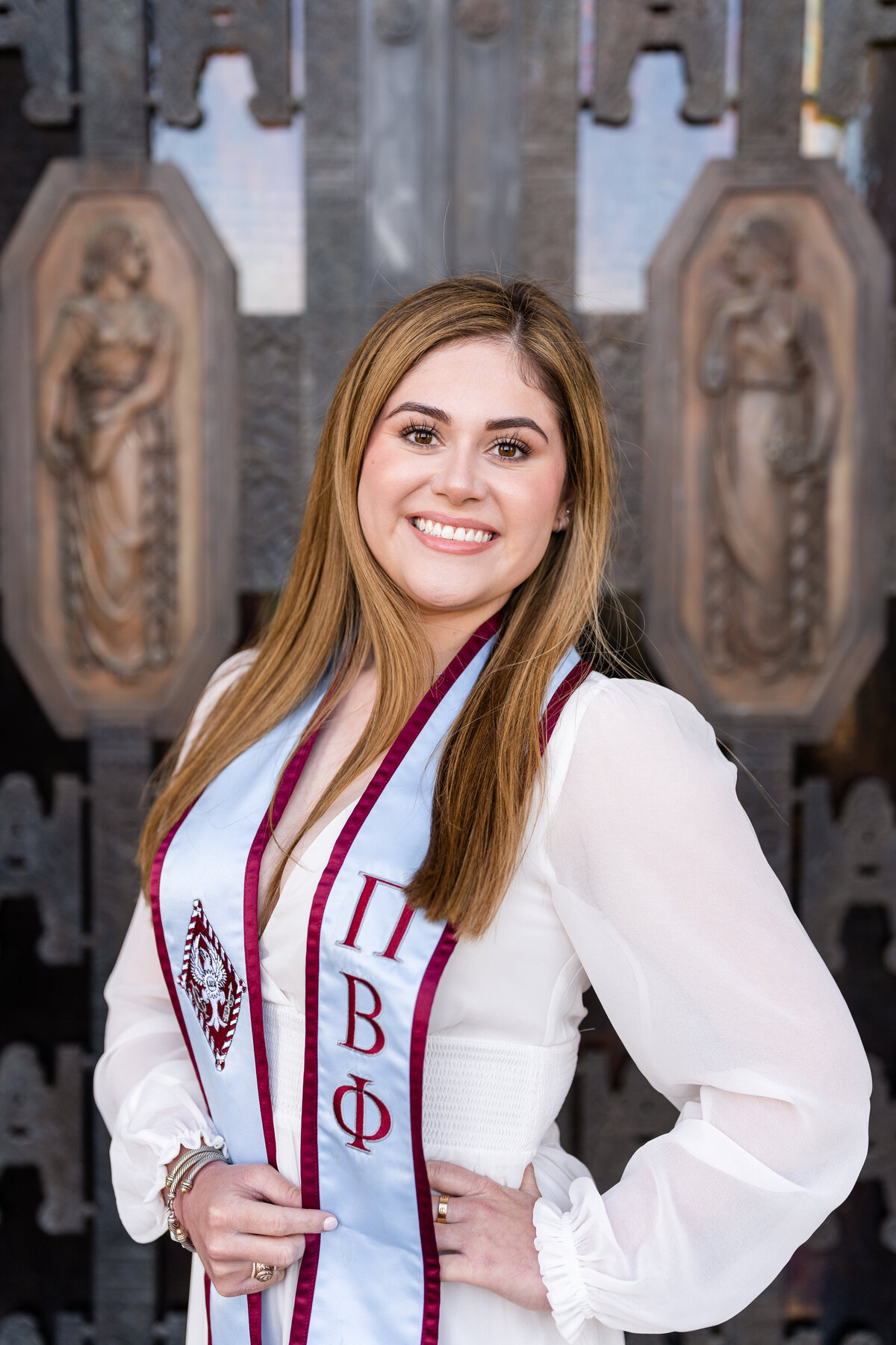 Texas A&M senior girl smiling with hand on hip and standing in front of Administration Building front doors  while wearing Pi Phi sorority stole and white dress