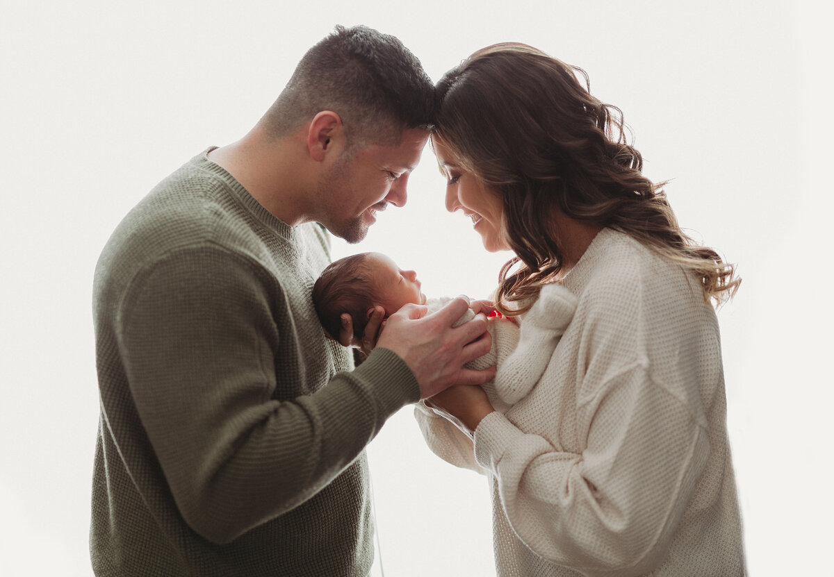 mom and dad holding baby in the middle on white backdrop with foreheads together