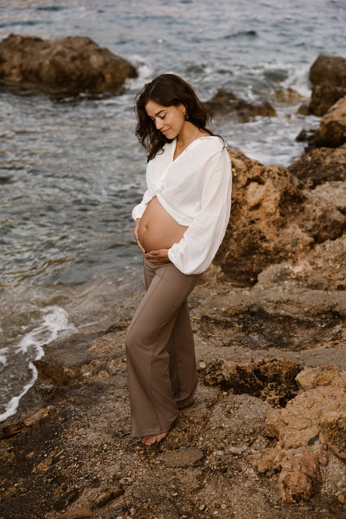 Woman is posing with her belly during a pregnancy shoot at a rocky beach on Ibiza