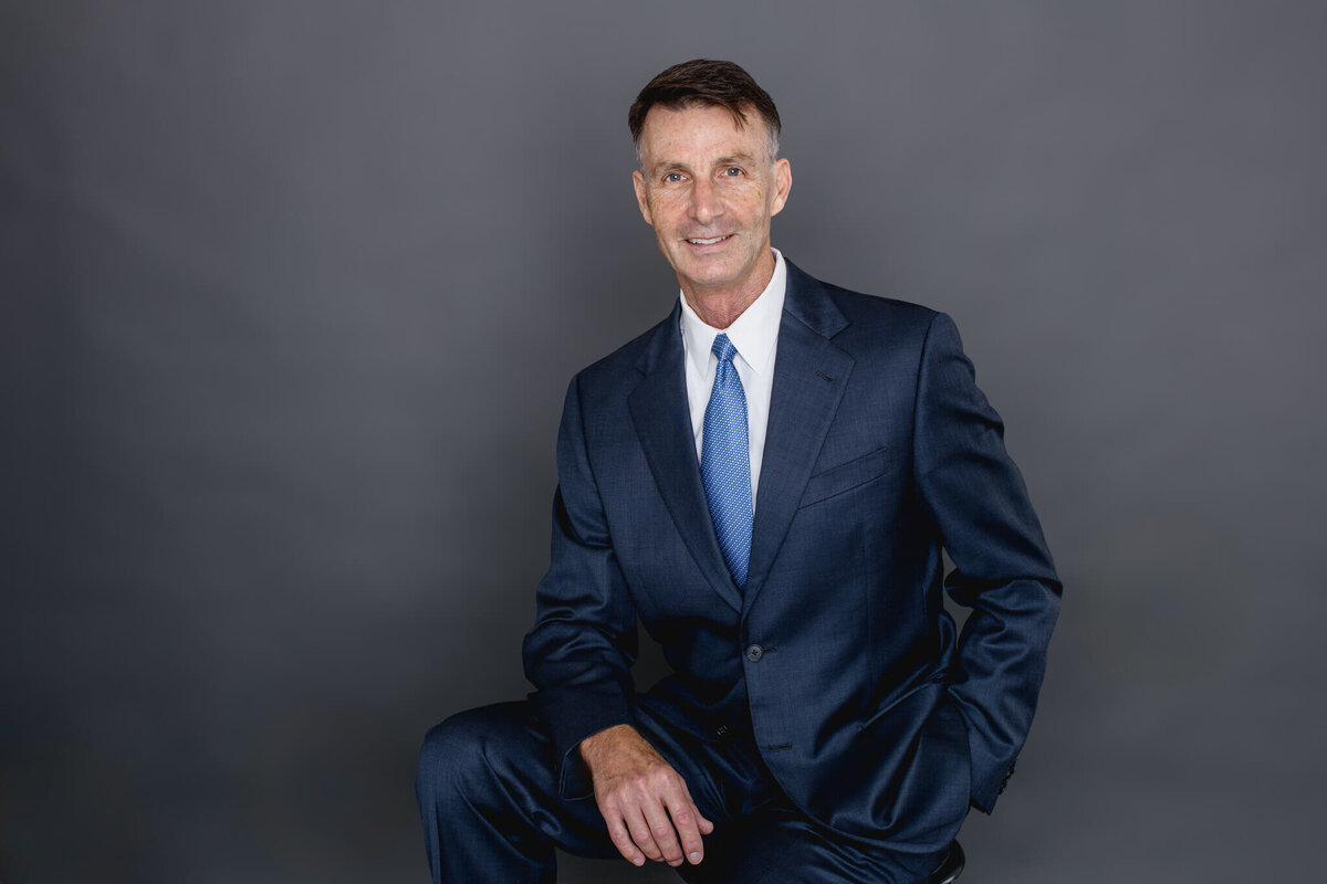 An older man with dark hair, wearing a navy blue suit jacket and light blue tie, looking at the camera with his right arm draped over his right thigh while sitting on a stool in front of a grey background in a Tustin studio.