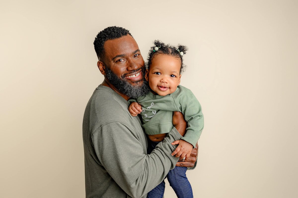 daughter is held by dad next to his face on a cream background wearing greens