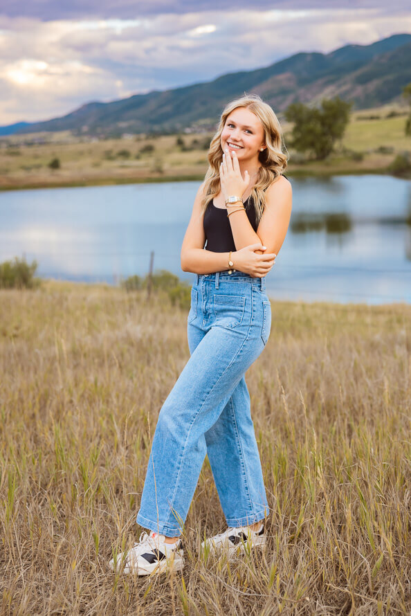 Girl in blue jeans laughs with lake and mountain in the background.