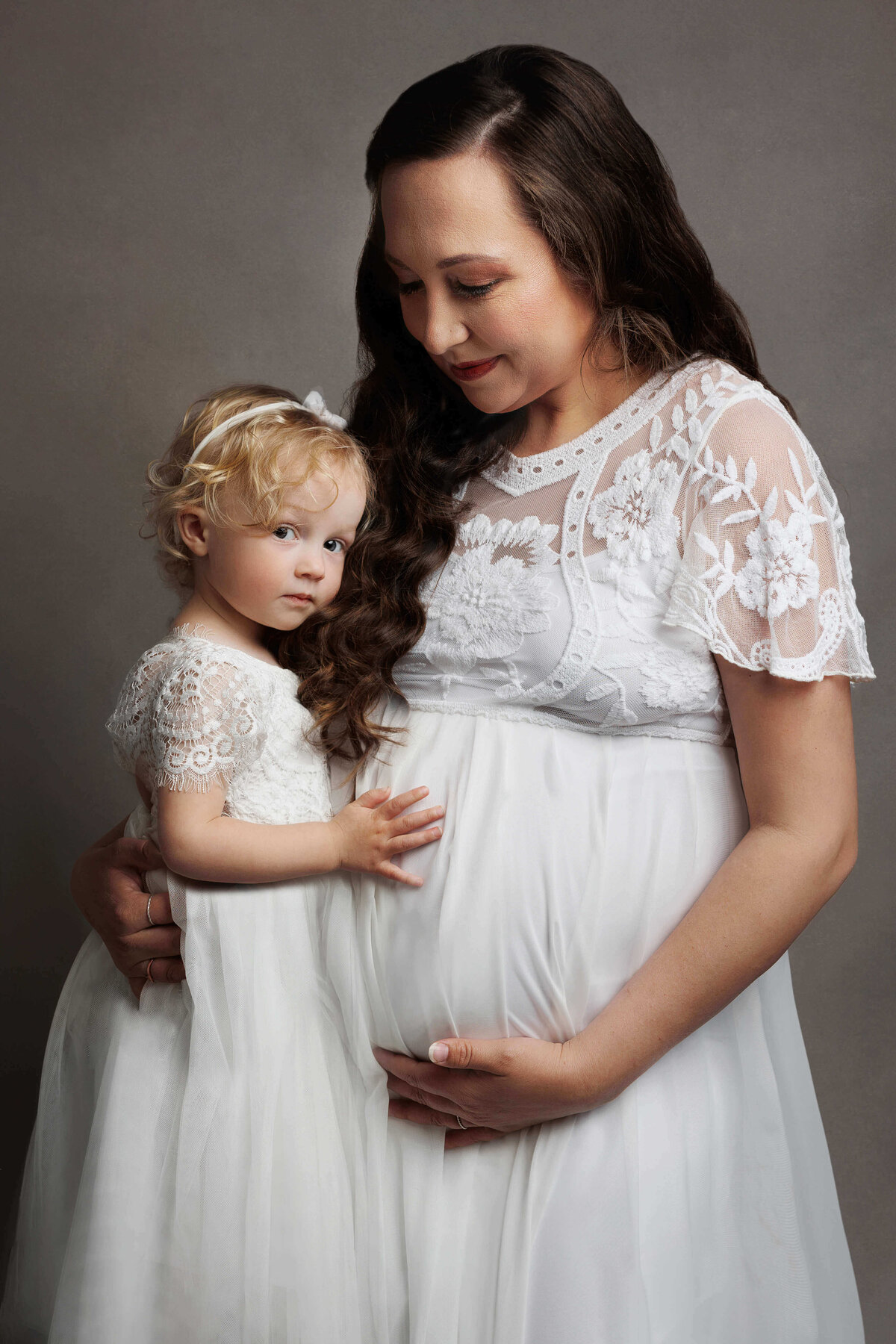 mother and daughter holding her pregnant belly wearing white dresses at a pregnancy photoshoot