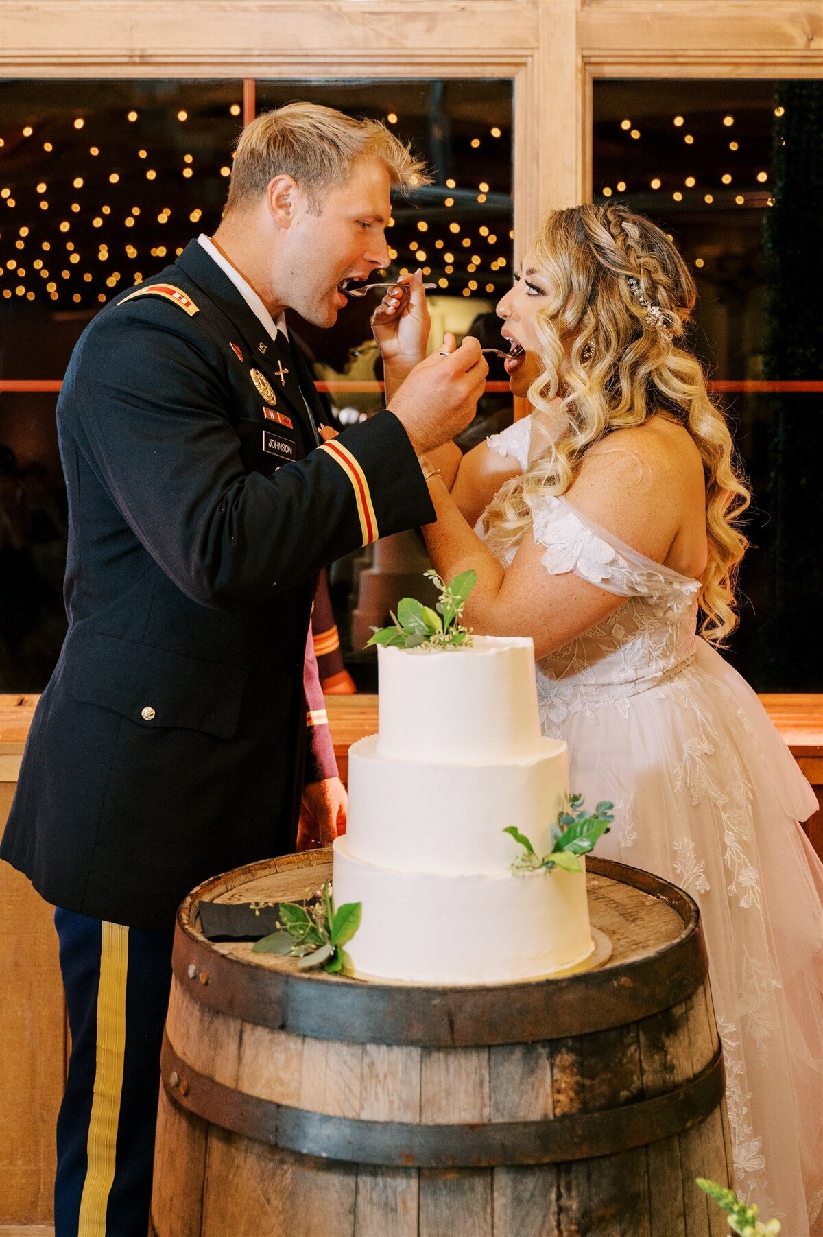 A man in a military uniform and a woman in a wedding dress joyfully feed each other cake at their Illinois wedding reception, with string lights in the background.