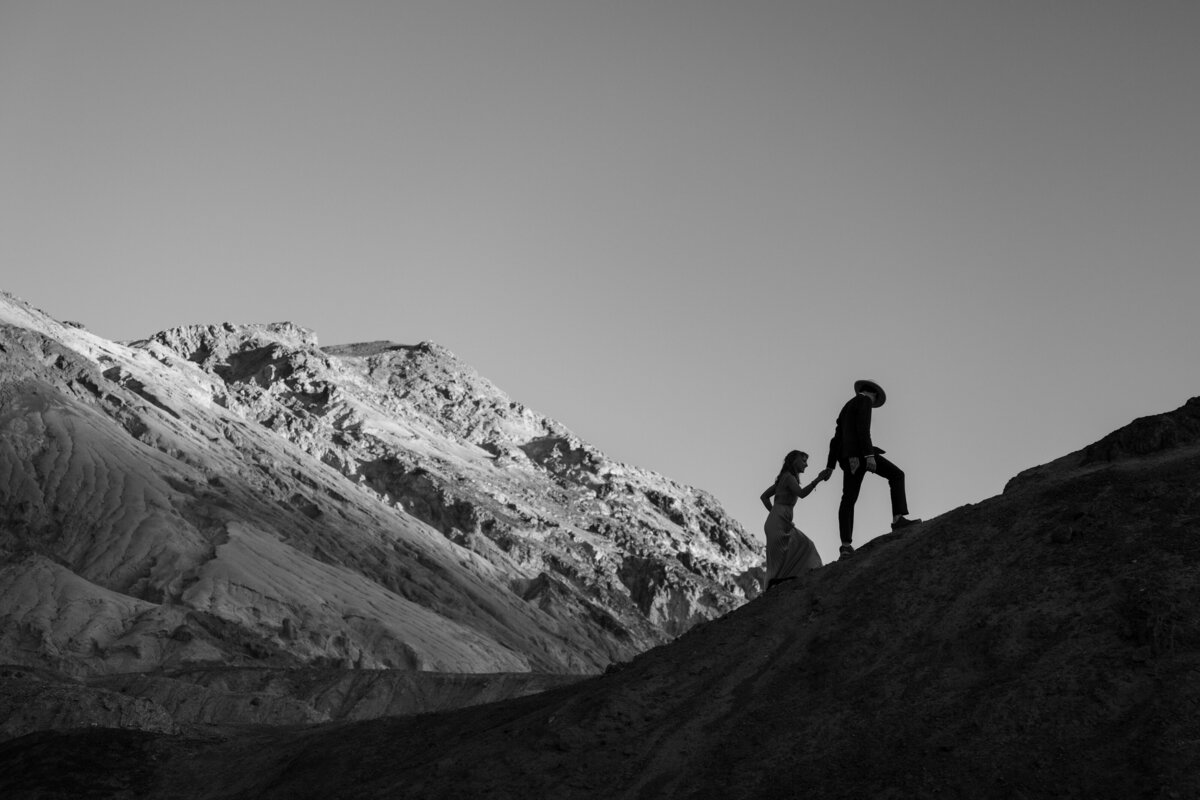 A couple holding hands and walking up the side of a rock